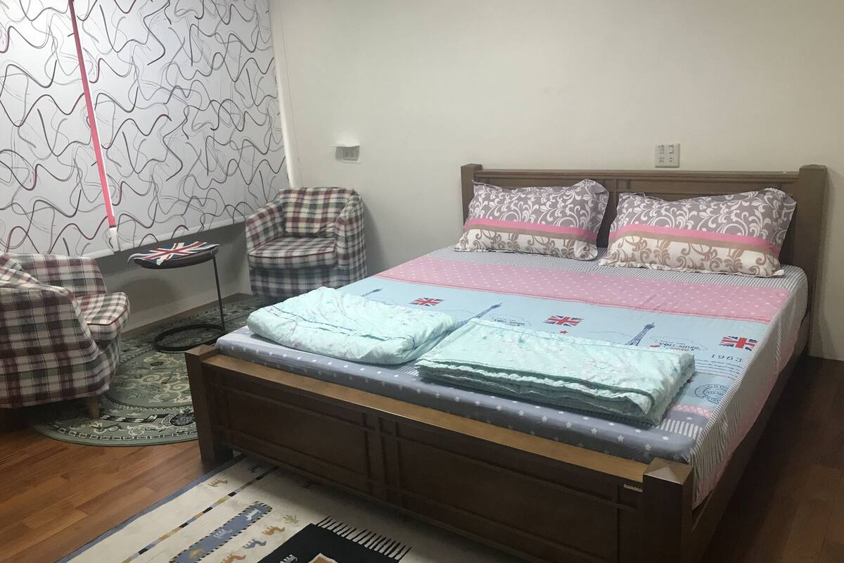 A bedroom with a wooden bed, a plaid armchair, a small table, and a patterned rug.