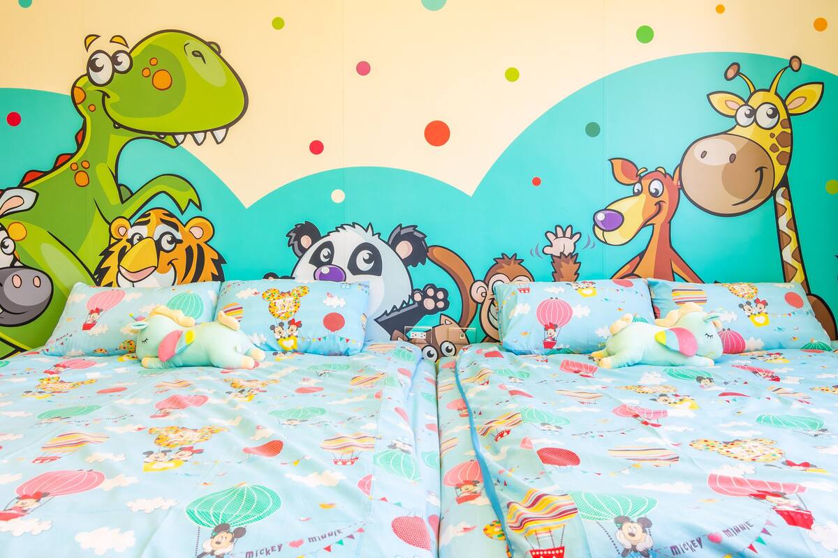 Una habitación infantil con una cama que tiene una colcha con temática de Mickey Mouse, almohadas con personajes de dibujos animados y un mural de varios animales en la pared.