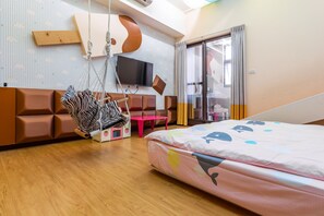 Ein modernes Hotelzimmer mit einer Couch, einem Hängekorbsessel, einem Flachbildfernseher, einem Bett mit farbenfroher Tagesdecke und einer Schiebeglastür nach draußen.