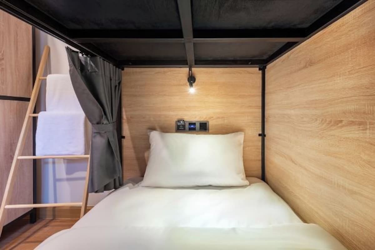 A compact, modern room with a wooden wall, a bed with white bedding, a ladder, and a small light fixture.