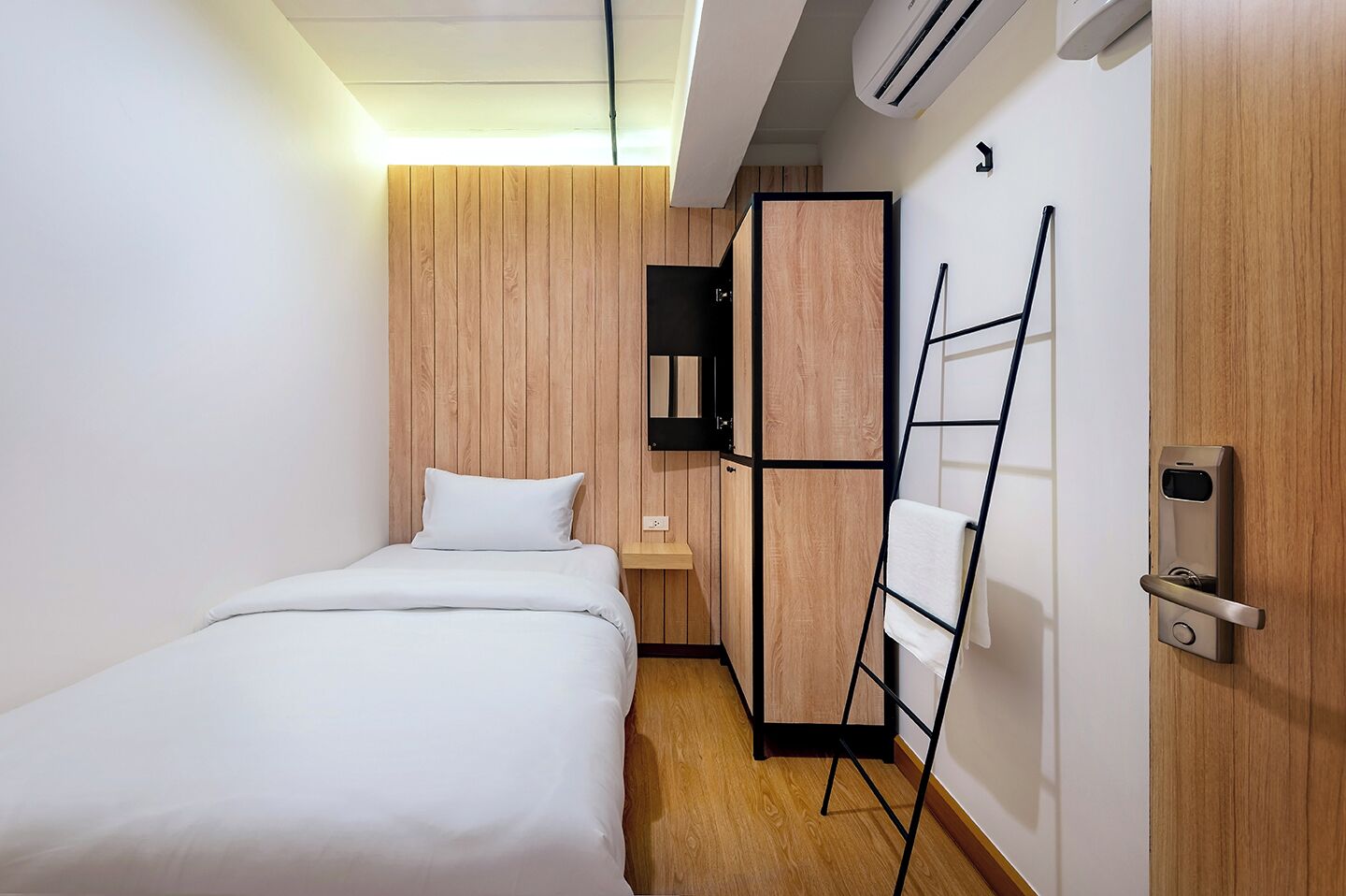 Una habitación de hotel pequeña y moderna con una cama, una mesita de noche, un armario y aire acondicionado integrado.