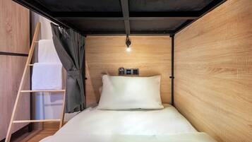 Een kleine kamer met een bed, een houten garderobe, een trapje en een klein plankje met een lamp.