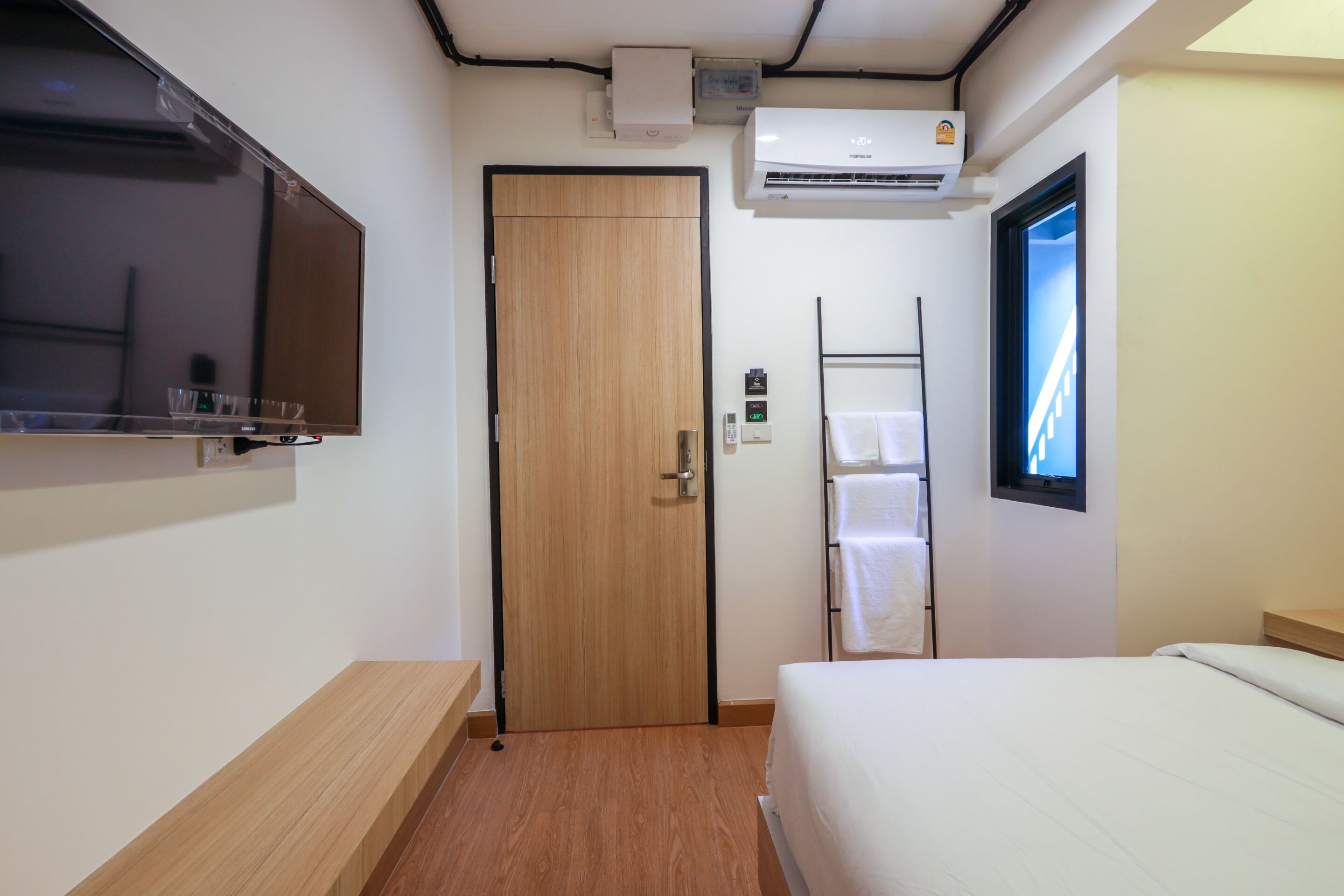 Una habitación de hotel con puerta de madera, televisor montado, unidad de aire acondicionado, toallero y cama.