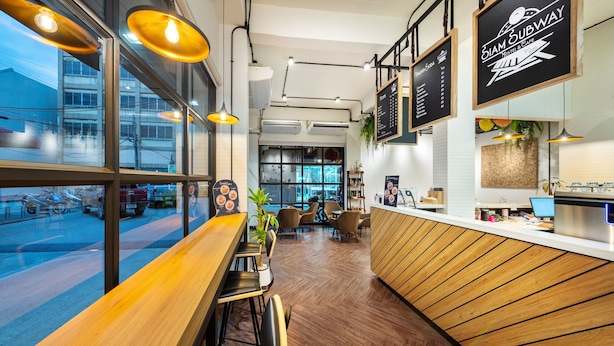 L’intérieur d’un café moderne, avec des meubles en bois, un comptoir où sont affichés les menus et de grandes fenêtres donnant sur la rue.