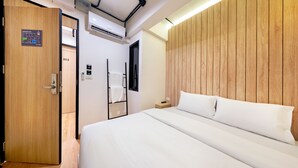 Une chambre d’hôtel avec un mur recouvert de panneaux de bois, un lit avec une literie blanche, un panneau indiquant la sortie de secours, un thermostat numérique et une porte dotée d’une poignée.