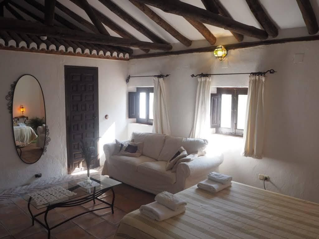 Una habitación acogedora con un sofá blanco, una mesa de vidrio y piso de baldosas. Hay una puerta y una ventana con cortinas.