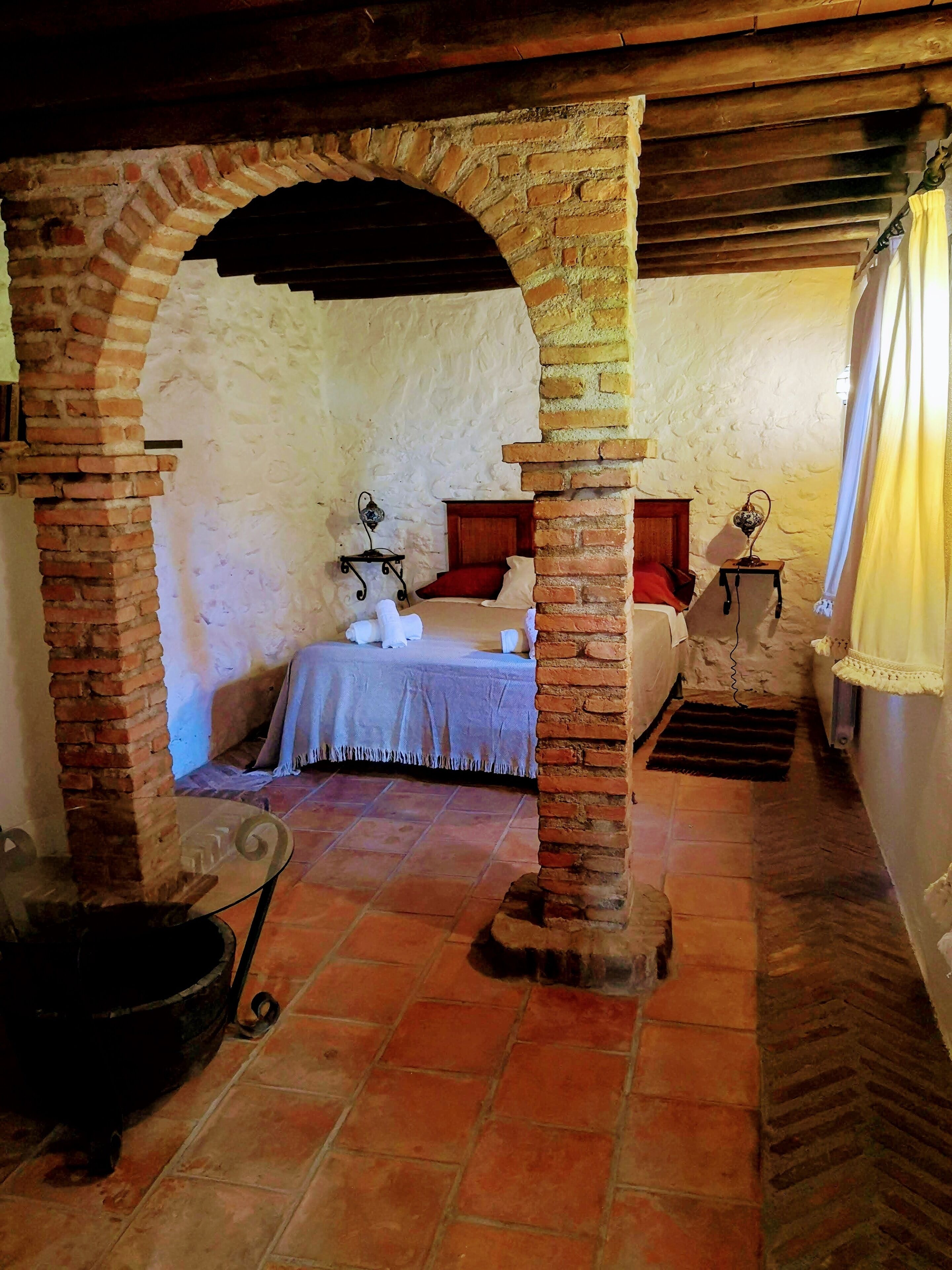 A rustic room with a brick archway, a bed with a red pillow, a small table with a lamp, and a window with a curtain.