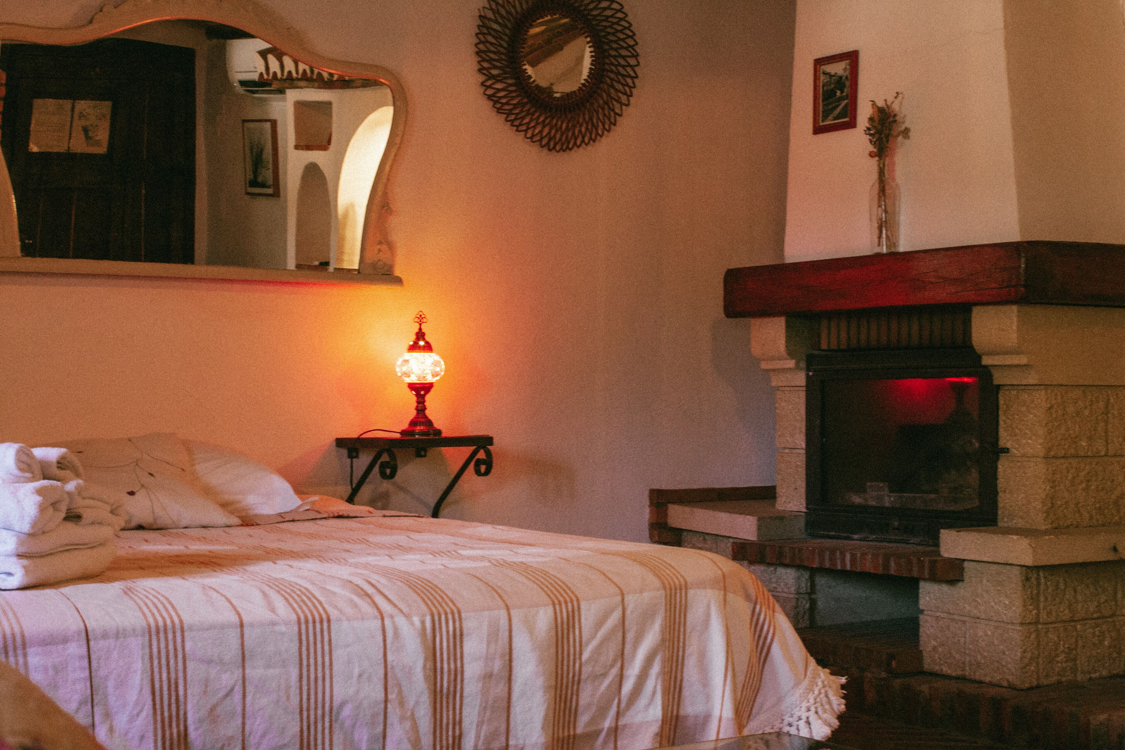 A bedroom with a bed, a bedside table with a lamp, a fireplace, and a mirror on the wall.
