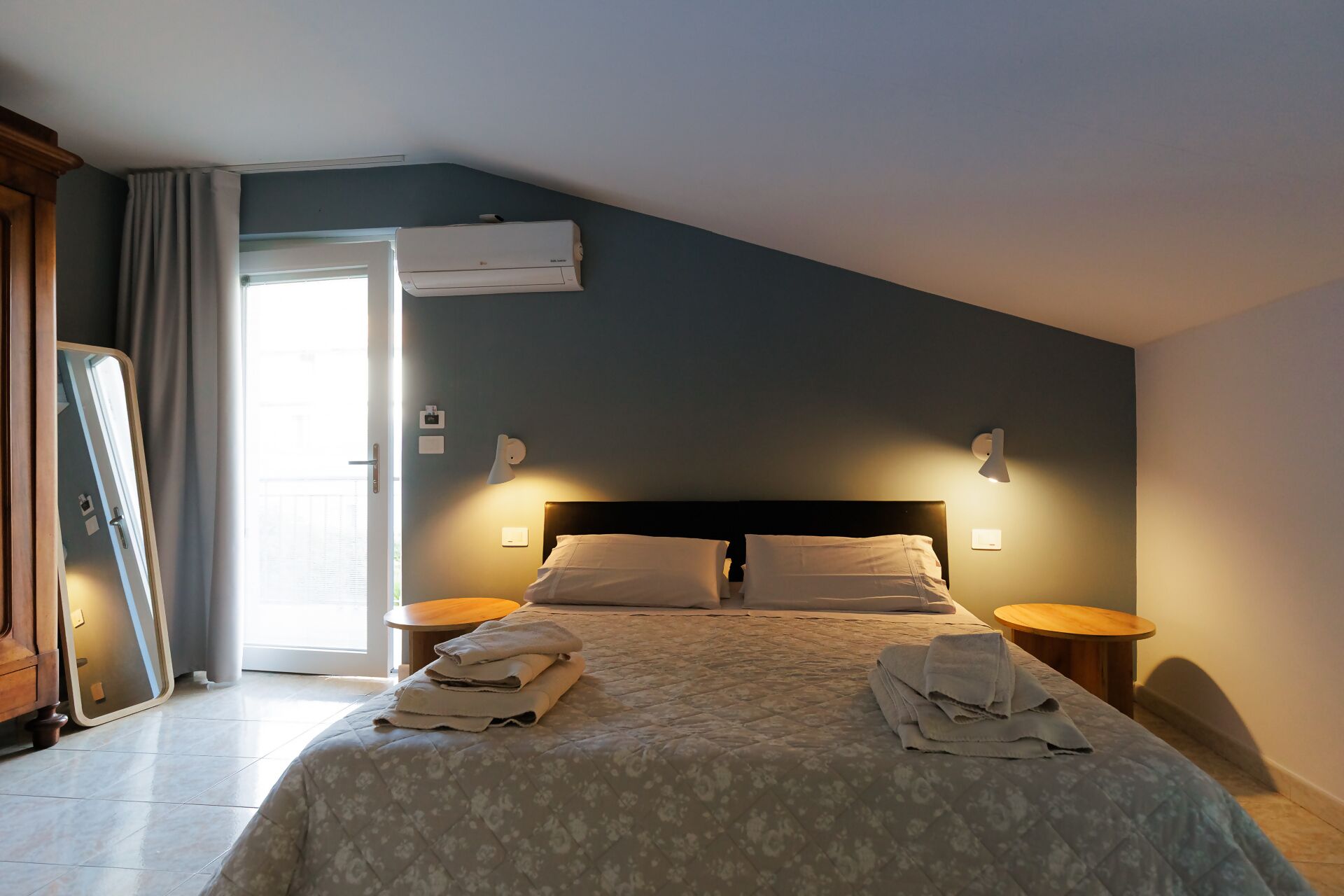 A bedroom with a bed, two bedside tables, a nightstand, a wall-mounted air conditioner, and a door leading to a balcony.