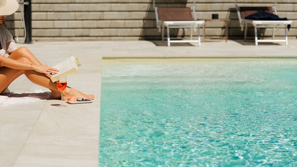 Eine Person sitzt am Pool, ein Buch und ein Glas Rotwein in der Hand.