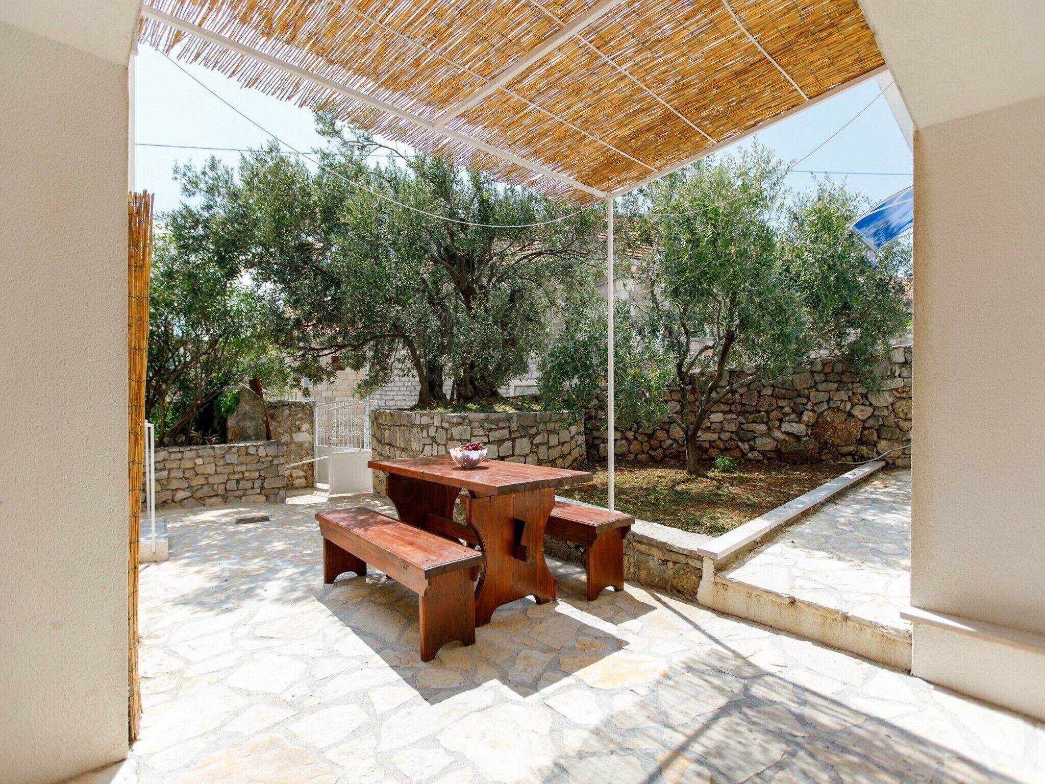 An outdoor patio with a wooden table and benches, surrounded by stone walls and trees.
