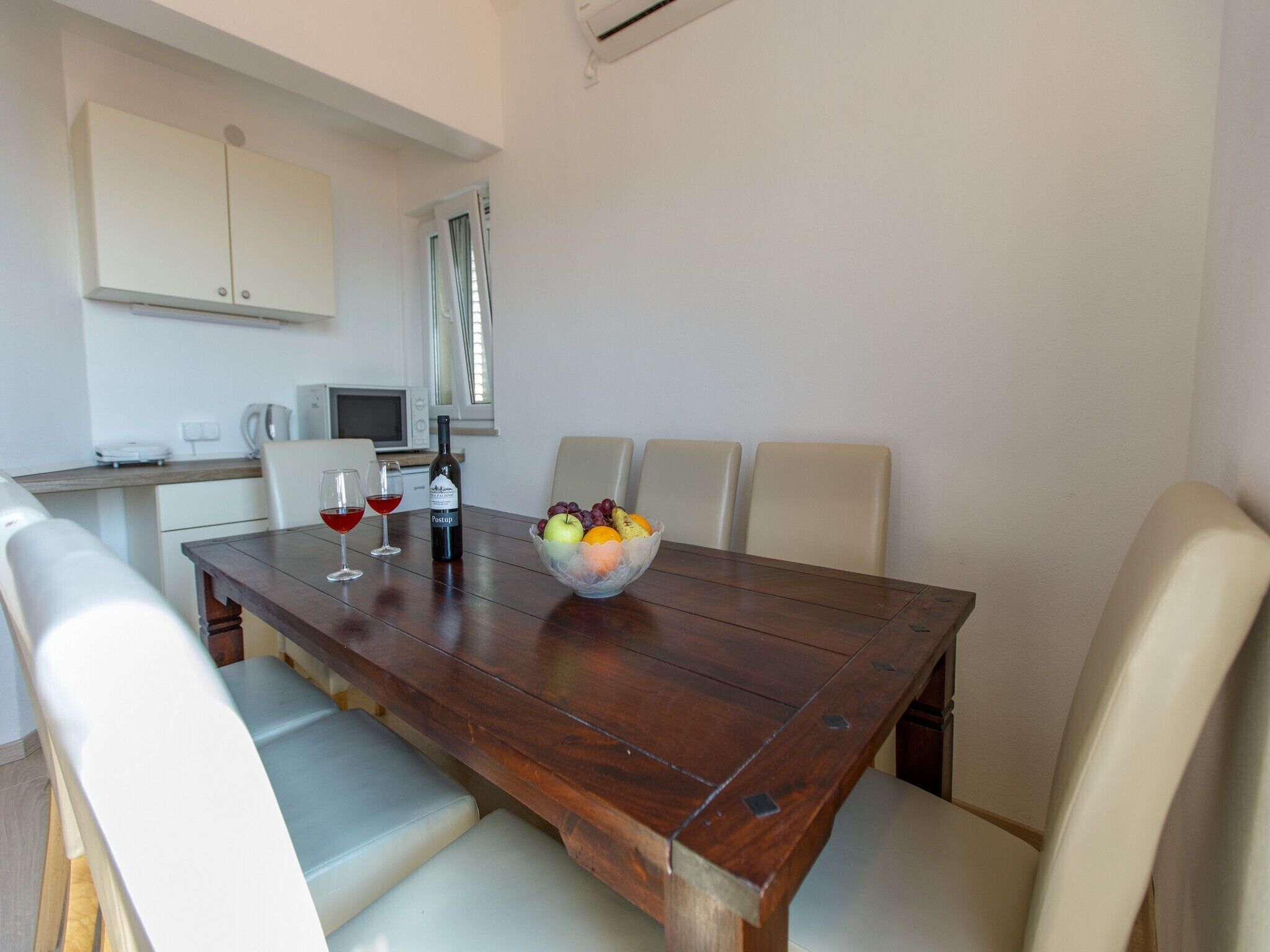 Un coin repas avec une table en bois, des chaises blanches, un saladier de fruits et deux verres de vin rouge.