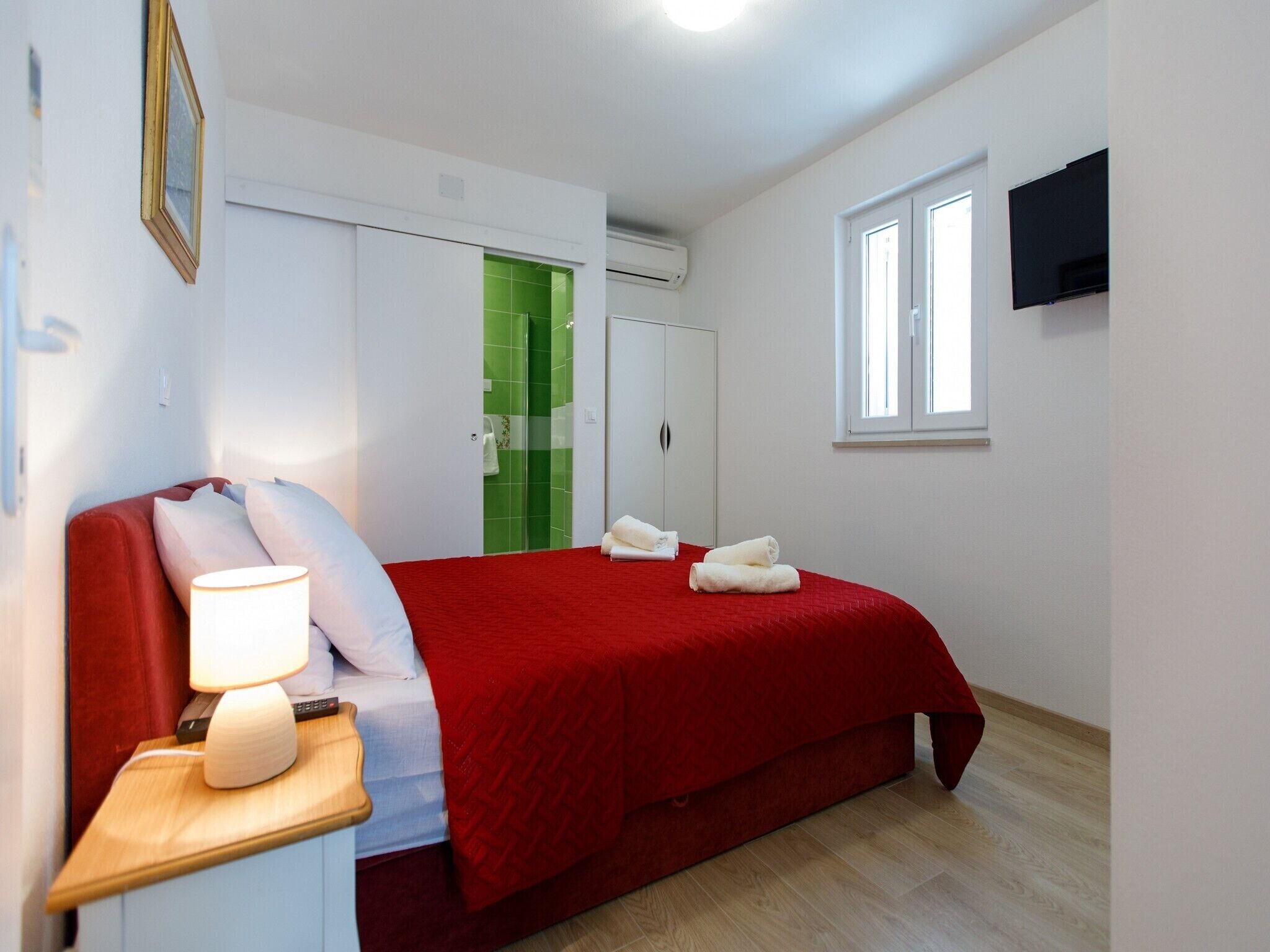 Et hotelværelse med seng, natbord, fjernsyn, garderobeskab og et bruseområde med grønne fliser.
