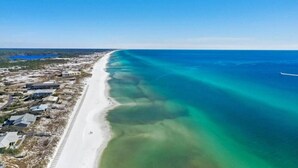 Une vue aérienne d’une zone côtière avec une longue plage de sable, des eaux d’un bleu limpide et quelques bâtiments dispersés.
