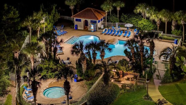 Un espace piscine avec des chaises longues et un petit bâtiment, le tout entouré de palmiers.