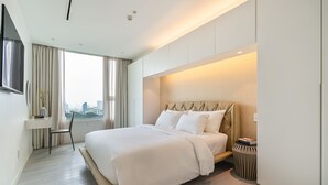 A modern hotel room with a large bed, a TV mounted on the wall, a desk with a chair, and a view of the city through large windows. - Saigon Finest - IDG Suites Collection - 12J (Ho Chi Minh City)