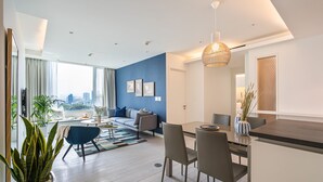 Een moderne woonkamer met een bank, eettafel en stoelen. Er is een groot raam met gordijnen, een potplant en uitzicht op de stad.