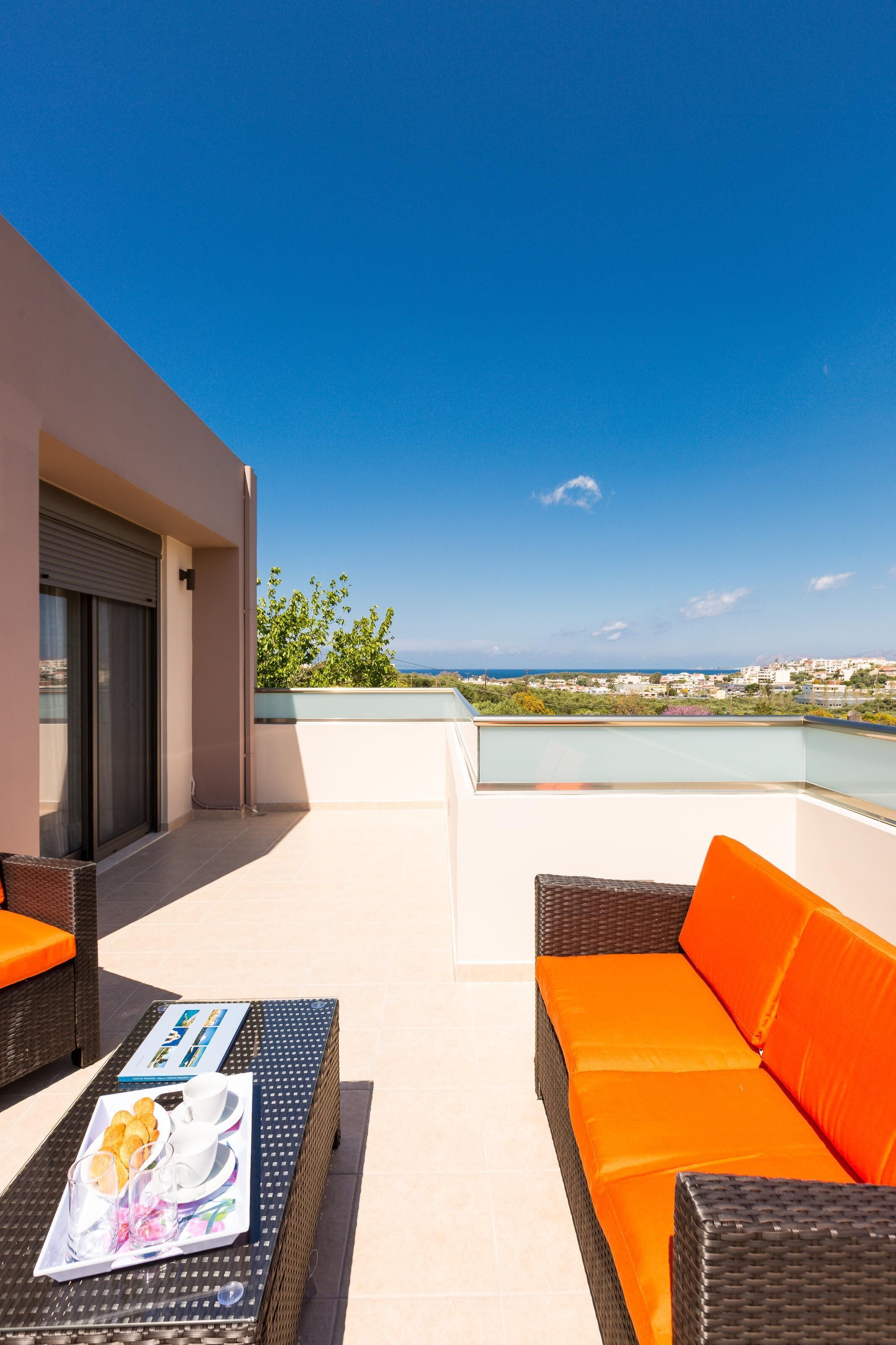 A balcony with a wicker sofa and table, a tray of food, and a view of the sea and cityscape.