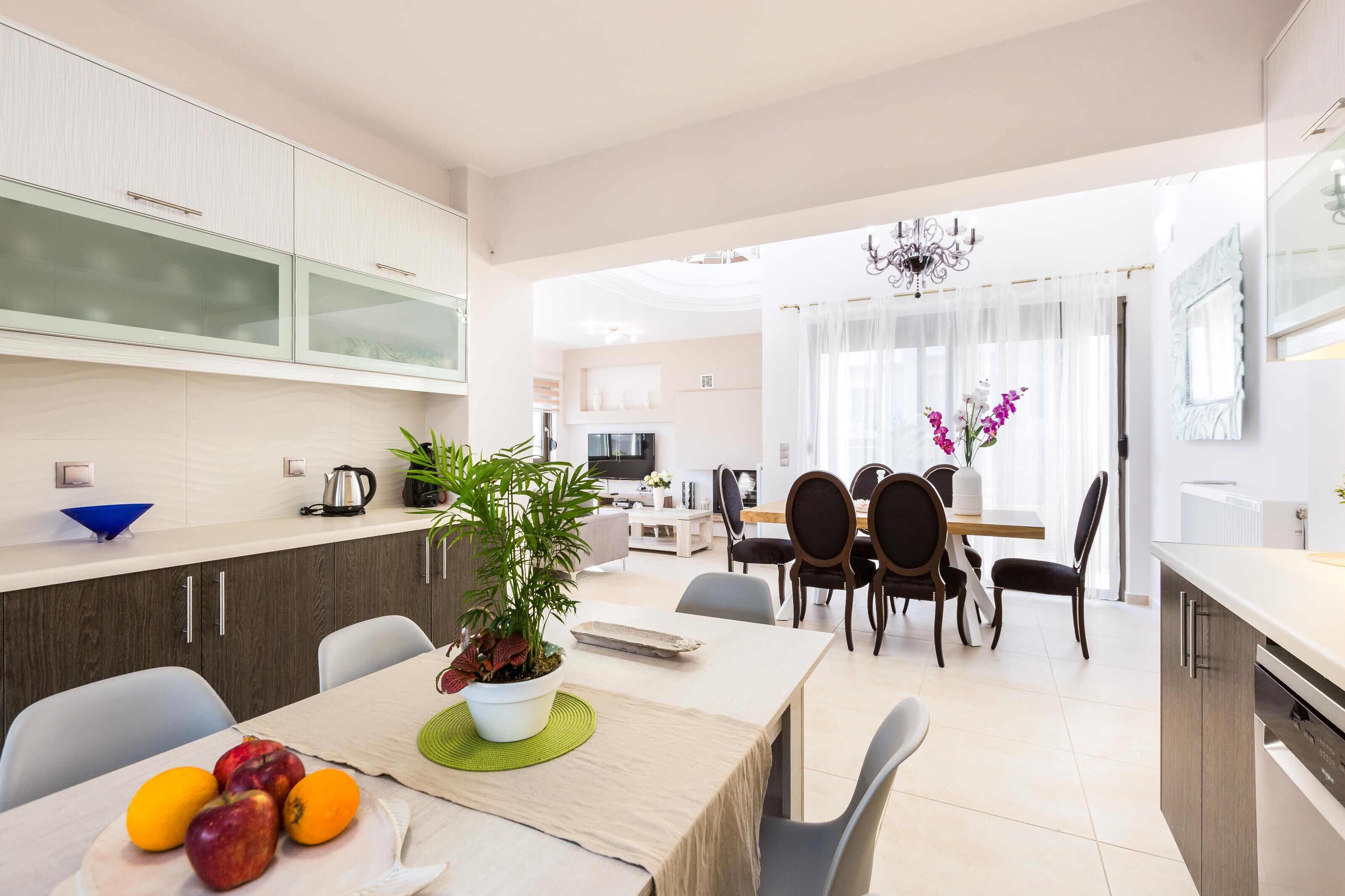 A modern kitchen with a dining area, featuring a table with fruits and a potted plant, and a view into the living room.