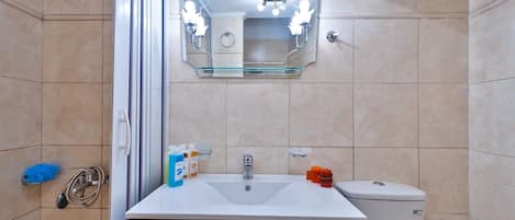 Un baño con un lavabo blanco, un espejo y un inodoro. Hay artículos de aseo en el lavabo y un cabezal de ducha en la pared.