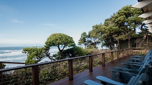 Una terraza de madera con mobiliario exterior, con vistas a una zona costera con árboles y el océano.