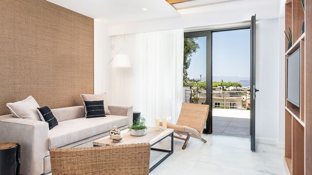Ein modernes Wohnzimmer mit einer Couch, einem Sessel, einem Couchtisch und Blick durch eine Glastür auf einen Balkon.