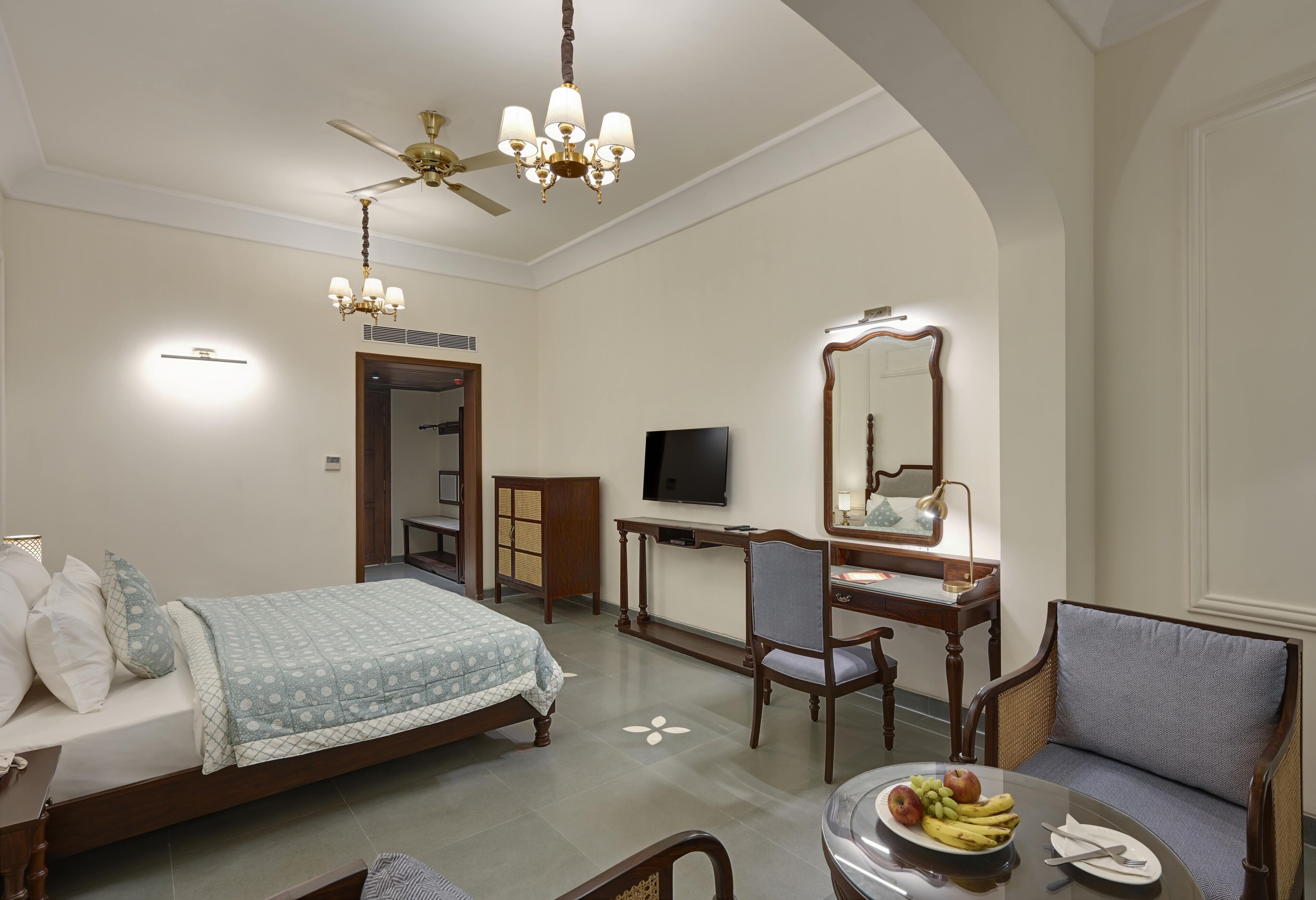 Quarto de hotel com uma cama, uma escrivaninha com televisão, uma cadeira, uma mesa de jantar com frutas e um candelabro.