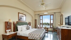 A hotel room with a large bed, a desk, a chair, a TV, and a view of a body of water. - Brahma Niwas - Luxury Lake View Hotel with Infinity Pool, Udaipur  (Udaipur)