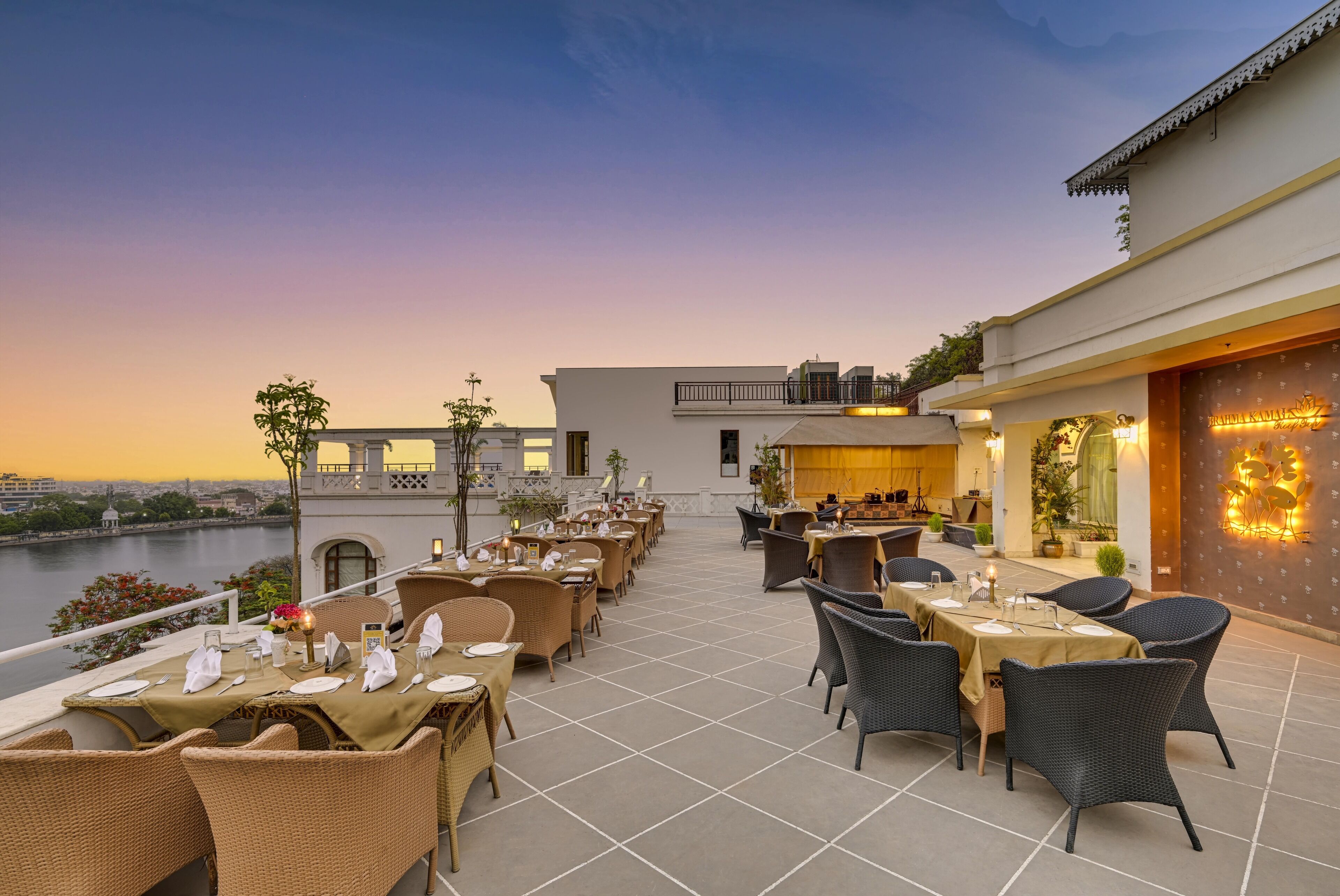 An outdoor dining area with tables and chairs, overlooking a body of water and a cityscape.