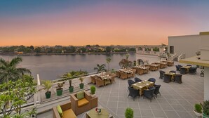 A terrace with outdoor seating, a view of a river, and a cityscape in the background. - Brahma Niwas - Luxury Lake View Hotel with Infinity Pool, Udaipur  (Udaipur)
