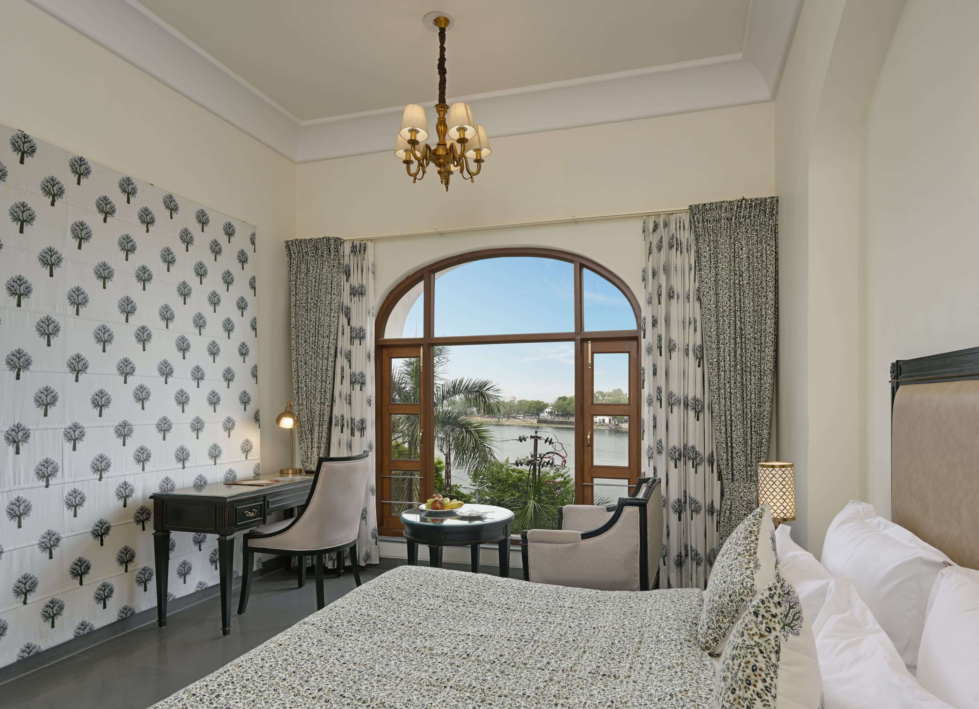 A hotel room with a large arched window, a desk with a chair, a round table with a lamp, and a bed with white bedding.