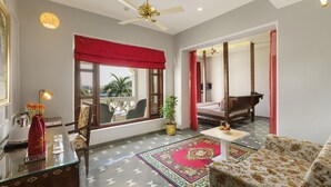 A spacious room with a large window, a ceiling fan, a red curtain, a sofa, a small table, and a decorative rug. - Brahma Niwas - Luxury Lake View Hotel with Infinity Pool, Udaipur  (Udaipur)