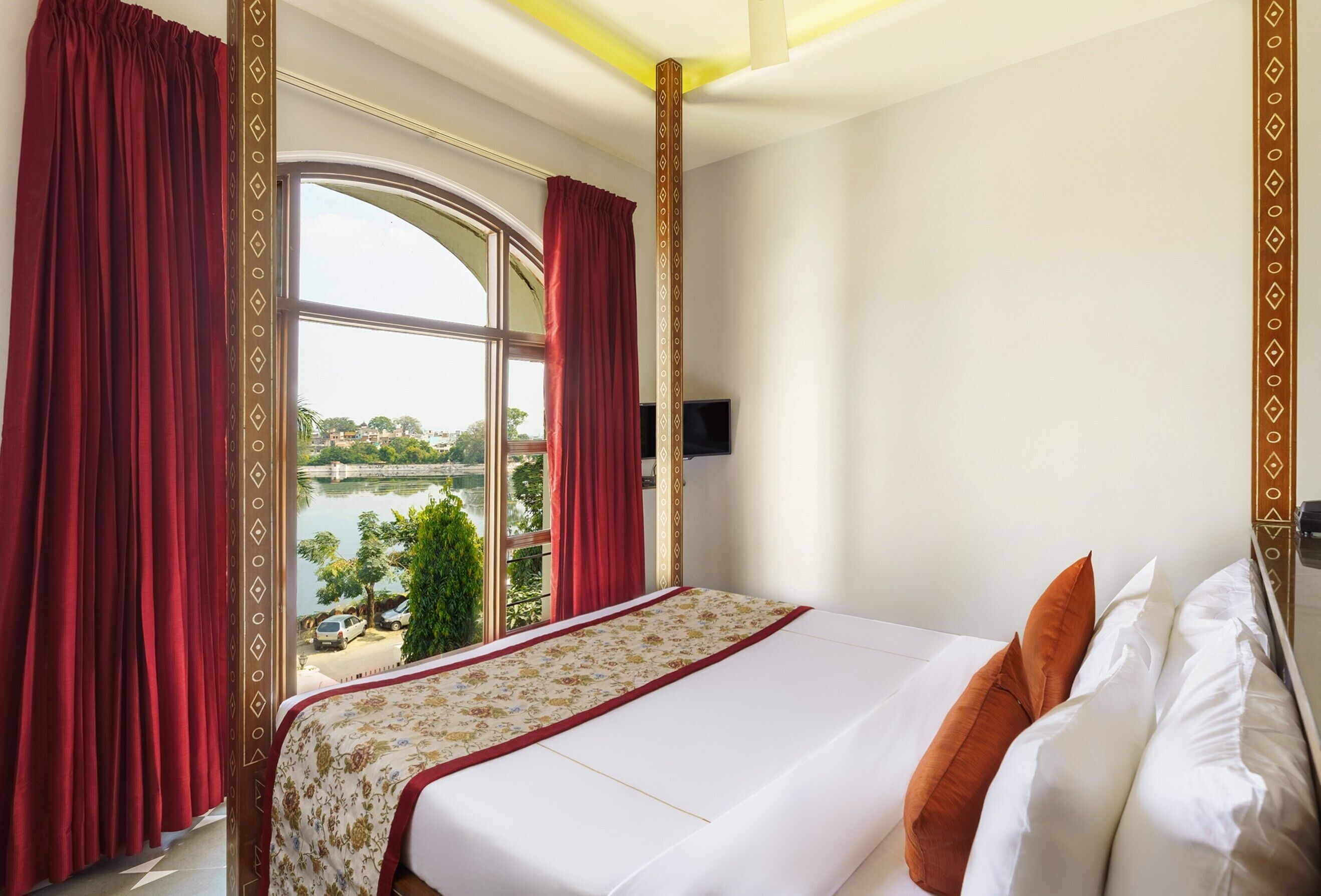 A hotel room with a large bed, red curtains, a view of a lake, and a TV mounted on the wall.