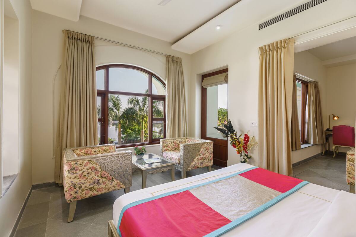 Una habitación de hotel con cama, una mesita, dos sillas y un ventanal con cortinas.