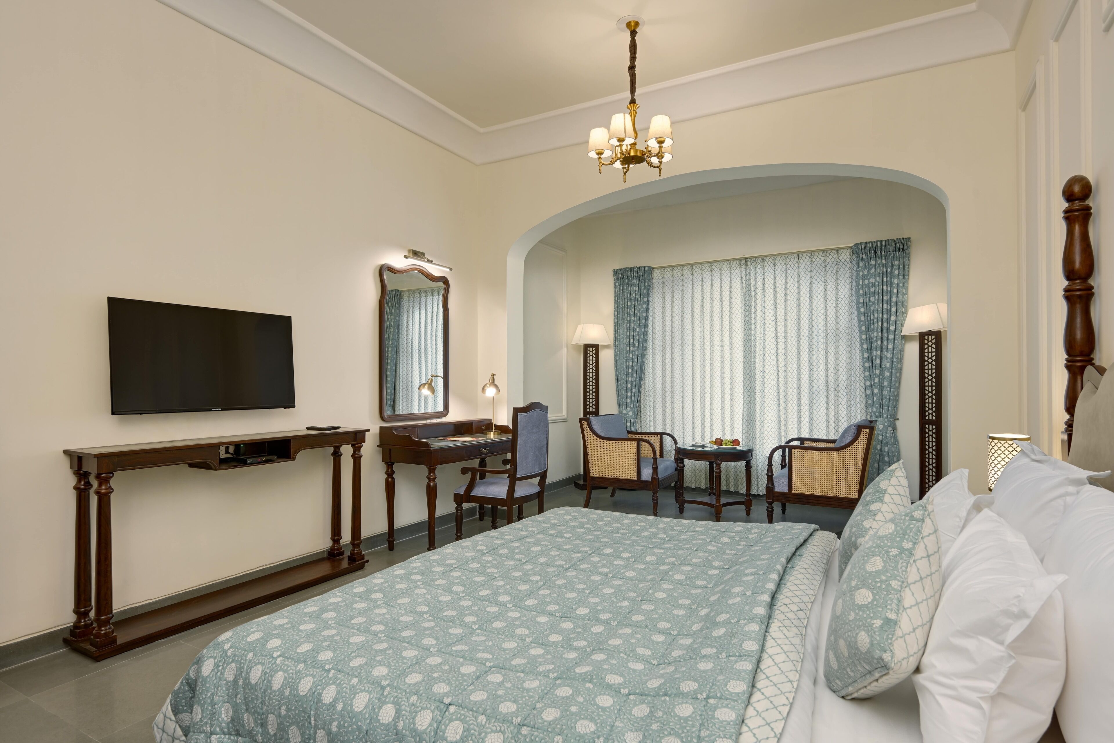 A hotel room with a large bed, a desk with a chair, a television, and a chandelier.