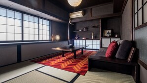 Una habitación tradicional japonesa con suelo de tatami, una mesa baja, un sofá con cojines, una lámpara colgante y vistas a través de paneles shoji.