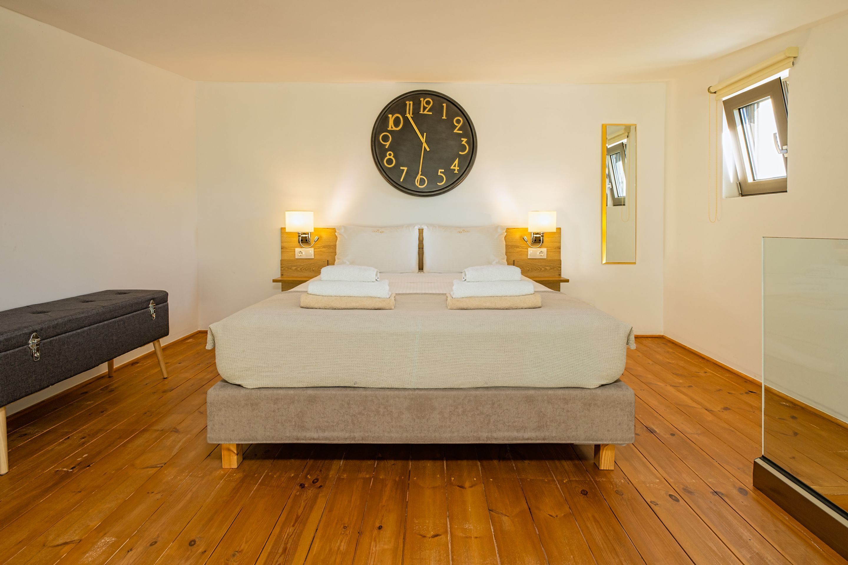 A bedroom with a large bed, wooden flooring, a wall clock, bedside lamps, a bench, and a window.