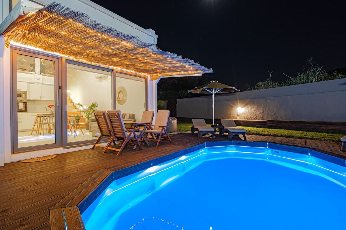 A swimming pool with wooden deck and lounge chairs, a covered patio area with string lights, and a view of the backyard.