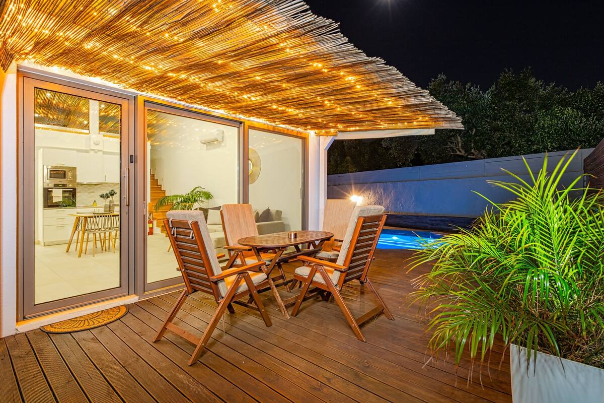 An outdoor deck with a wooden table and chairs, a glass door leading to a kitchen, and a pool in the background.