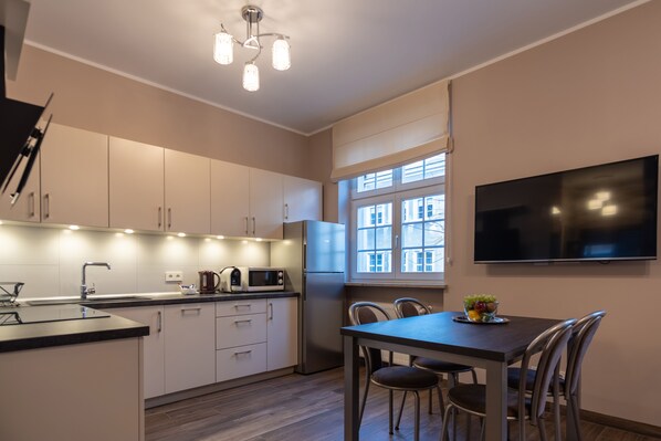 A modern kitchen with white cabinets, a built-in microwave, and a refrigerator. There is a dining table with chairs and a flat-screen TV mounted on the wall.