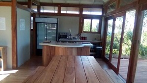 Une terrasse en bois avec une table centrale et des bancs, un coin cuisine équipé d’un four à micro-ondes et d’une cuisinière, et de grandes fenêtres offrant une vue sur la verdure.