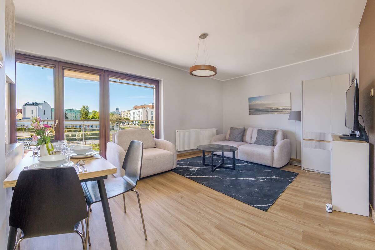 A modern living room with a dining area, large windows, a sofa, a coffee table, and a flat-screen TV.