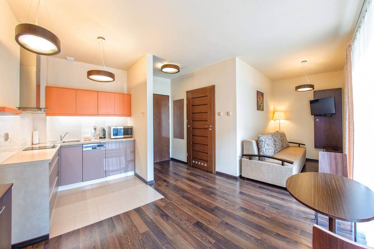 A modern kitchen with a built-in dishwasher, a dining area with a wooden table, and a living area with a sofa and a TV.