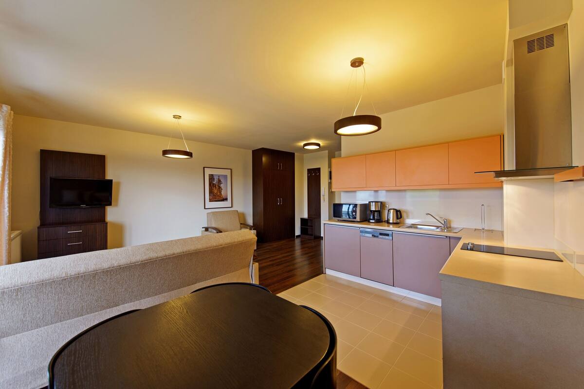 A modern kitchen with a microwave, coffee maker, and sink. There is a dining area with a table and chairs. The room has wooden flooring and a large window.