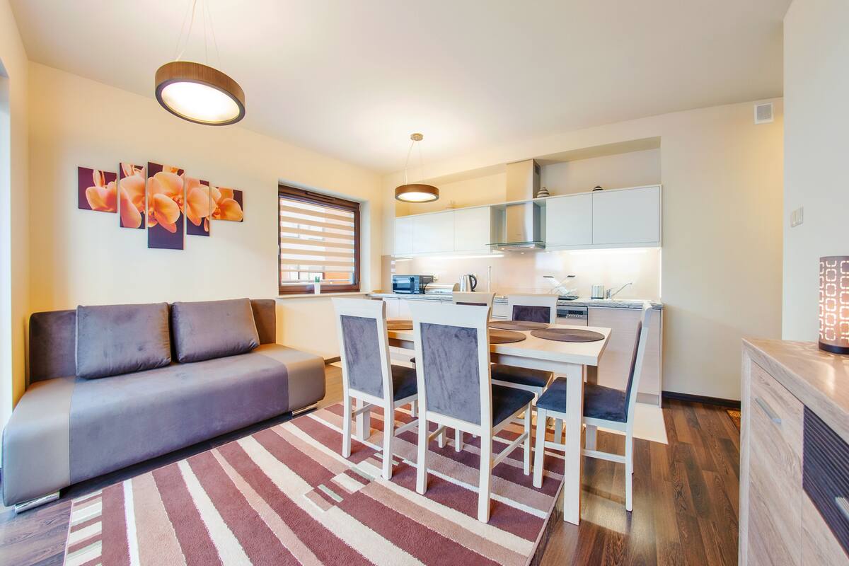 A modern living room with a sofa, dining table, and chairs. There is a kitchen area with cabinets and a countertop.