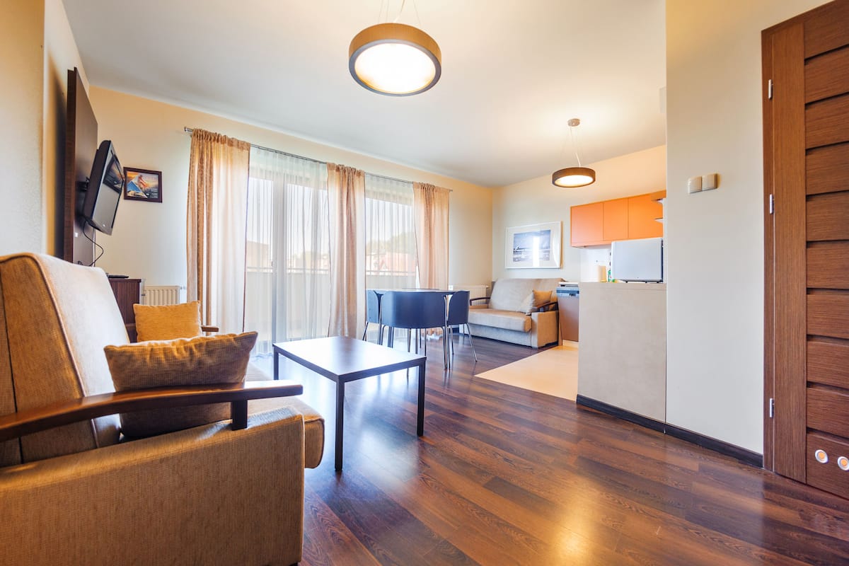 A modern living room with a sofa, a coffee table, and a TV mounted on the wall. There is a kitchen area with cabinets and a refrigerator in the background.