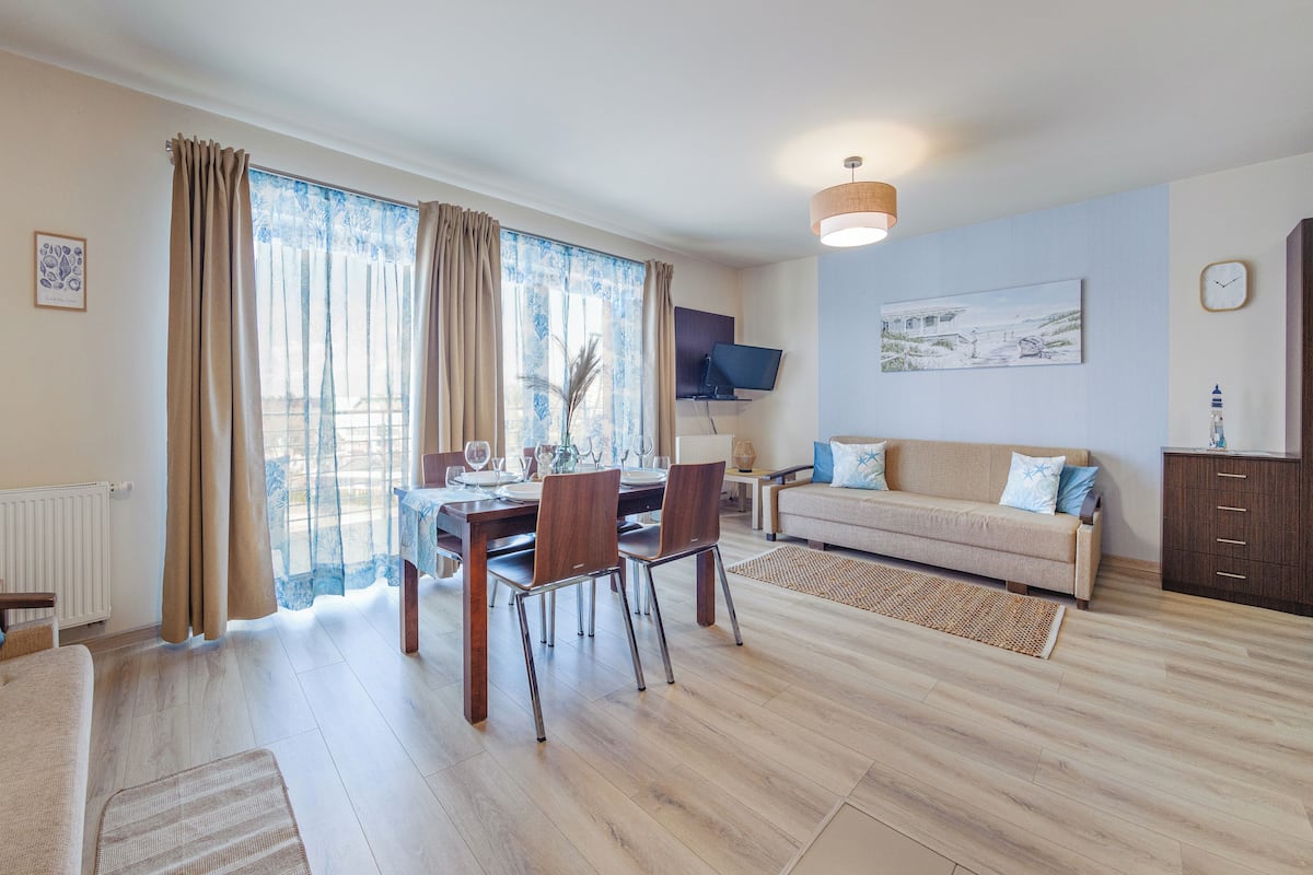 A modern living room with a dining area, a sofa, a TV mounted on the wall, and a clock.