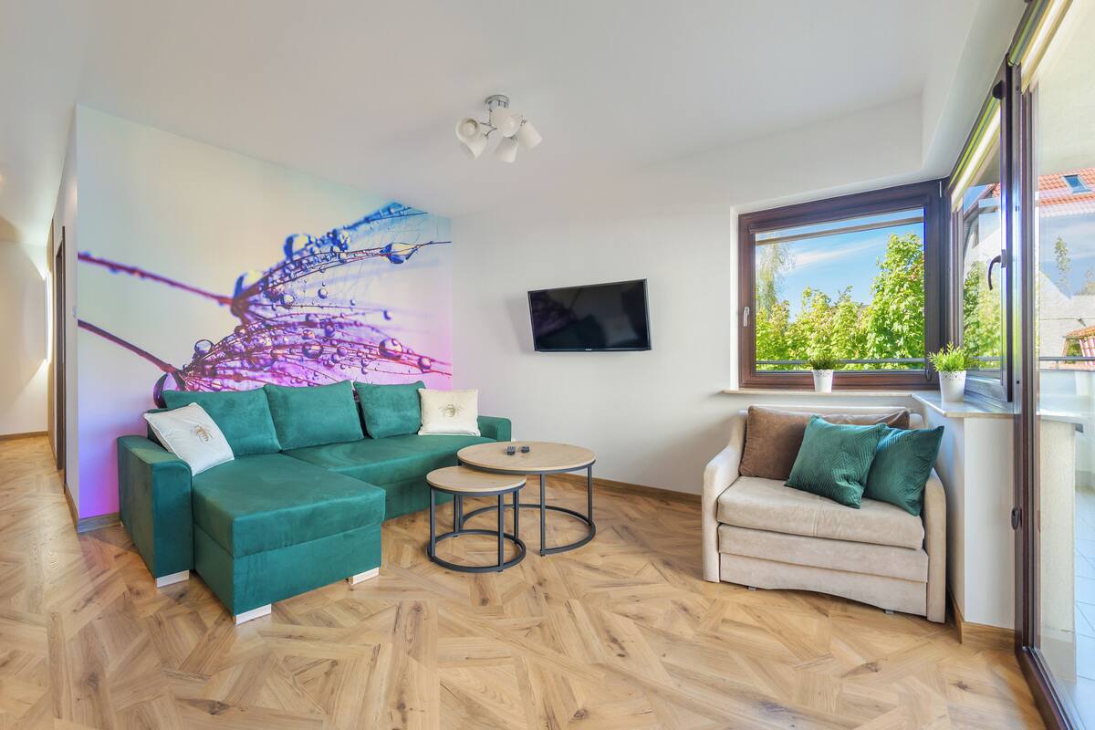 A modern living room with a green sectional sofa, a beige armchair, a small round table, and a large abstract painting on the wall.