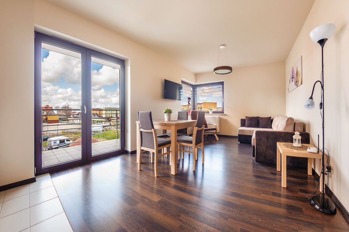 A spacious living area with a dining table and chairs, a sofa, a TV mounted on the wall, and a large window with a city view.