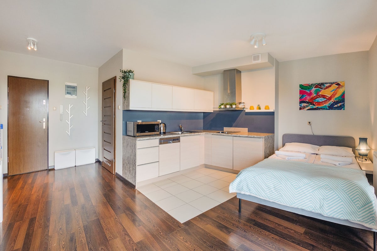 A modern hotel room with a kitchen area, a bed with a patterned bedspread, and a colorful abstract painting on the wall.